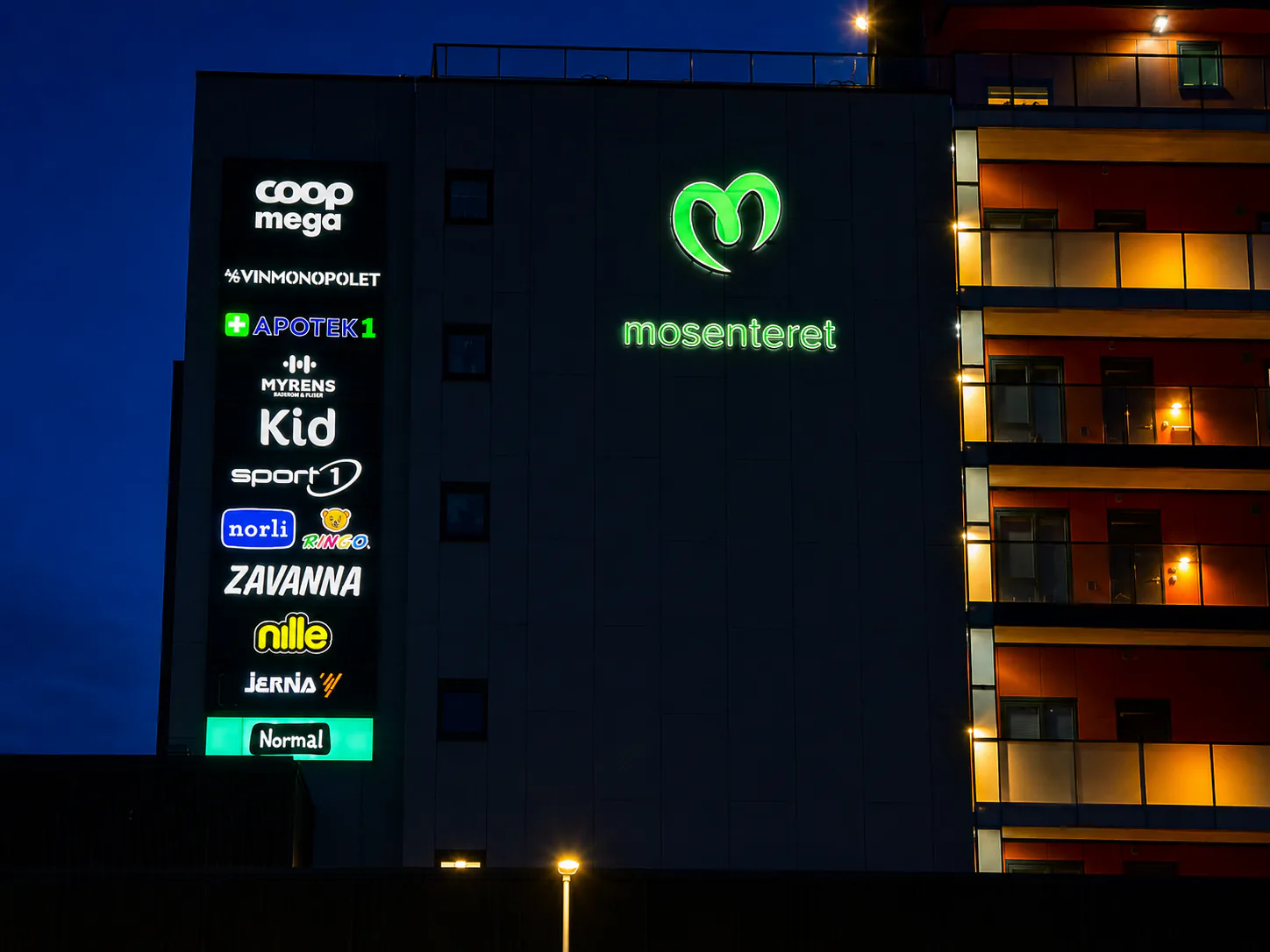 Mosenteret i grønt lys med opplyst butikkpylon på fasaden av et leilighetsbygg
